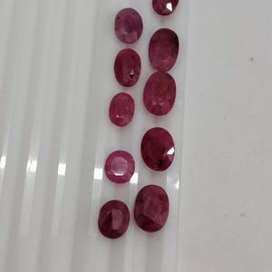 May include: A collection of nine loose, oval-cut rubies in varying shades of deep red and pink. The gemstones are arranged in two vertical columns on a white, ridged surface, showcasing faceted surfaces that reflect light across their polished tops.