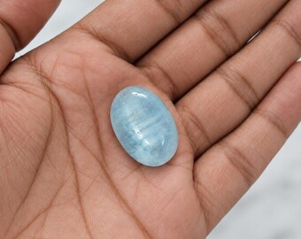 May include: An oval-shaped, polished gemstone with a translucent, pale blue color and subtle internal horizontal banding. The stone features a smooth, rounded surface and a glossy finish, displaying variations in light and dark blue tones throughout.