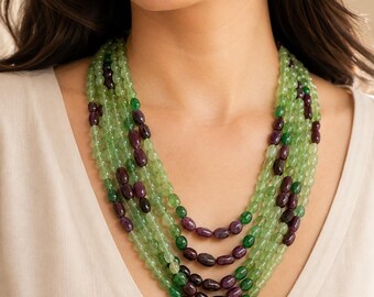 May include: A multi-strand necklace featuring polished light green beads interspersed with dark purple oval beads. The necklace consists of several layered strands of varying lengths, creating a full, textured appearance around the neck area.