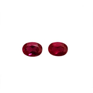 May include: Two oval-shaped, faceted red gemstones with a brilliant cut. The stones exhibit a deep, saturated crimson hue and a reflective surface with multiple geometric facets that catch the light, creating a clear, polished appearance.