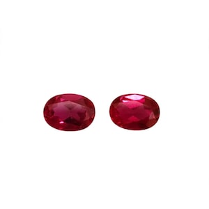 May include: Two oval-cut, deep red gemstones with a faceted surface. The stones are translucent and display a vibrant, saturated crimson hue with light reflecting off the polished, angled surfaces of the cut gems against a white background.
