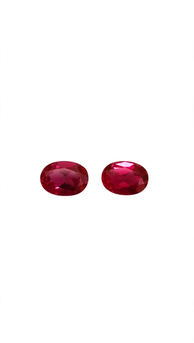 May include: Two oval-cut, deep red gemstones with a faceted surface. The stones are translucent and display a vibrant, saturated crimson hue with light reflecting off the polished, angled surfaces of the cut gems against a white background.