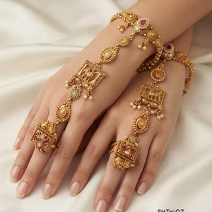 May include: A pair of gold-toned hand harnesses featuring intricate filigree work, square pendants with embossed elephant motifs, and small red stones. The jewellery connects a wrist bracelet to a ring on the middle finger with decorative links and small white beads.