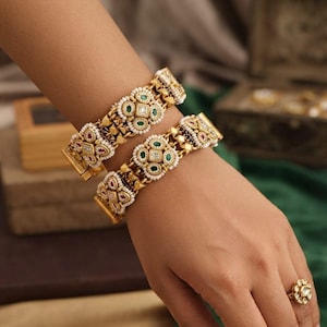 Op de afbeelding: Twee goudkleurige armbanden met ingewikkeld metaalwerk, kleine witte zaadparels en ingelegde groene en rode stenen. De armbanden hebben een breed, gestructureerd ontwerp met herhalende bloemmotieven. Een bijpassende goudkleurige ring met een centrale steen en omringende bloemblaadjes is ook zichtbaar.