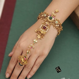 May include: A gold-toned hand harness featuring a central square red stone, green accents, and intricate floral filigree. The piece includes a linked chain connecting the wrist cuff to a ring adorned with a detailed embossed motif. Text reads: No:-235. HERITAGE PANJA D:-D-85.