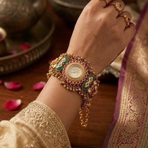 Puede incluir: Un brazalete reloj dorado con una esfera blanca redonda rodeada de pequeñas piedras rojas y detalles en esmalte verde. La pieza incluye intrincados adornos de cuentas doradas y una cadena delicada, junto con tres anillos dorados a juego.