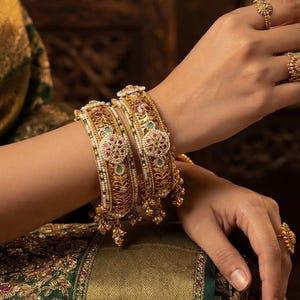 May include: Two gold-toned bangles featuring intricate filigree metalwork, embedded with small white pearls, red and green gemstones, and dangling gold charms. The jewelry set includes matching gold rings with similar ornate floral-inspired detailing.