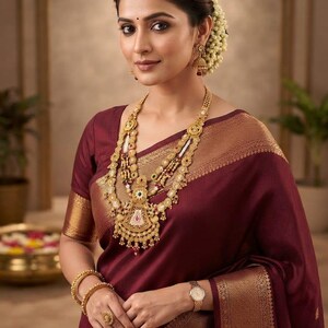 May include: A deep maroon saree with a wide gold border, paired with an elaborate multi-layered gold necklace featuring intricate filigree, matching gold jhumka earrings, two gold bangles, a gold ring, and a gold-toned wristwatch.