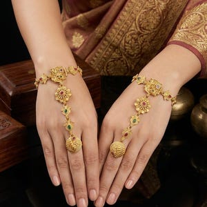 May include: A pair of gold-toned hand harnesses featuring intricate floral and leaf motifs. Each piece consists of a wrist bracelet connected by a delicate chain to a central flower element, ending in a ring with a circular textured medallion.