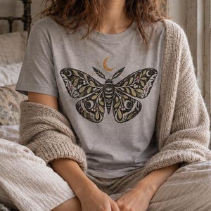 May include: A light gray short-sleeve t-shirt featuring a large, detailed graphic of a moth with intricate botanical and celestial patterns on the wings, including leaves, berries, and crescent moons, centered on the chest area.