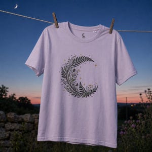 Könnte beinhalten: Ein hellviolettes Kurzarm-T-Shirt mit einer zentralen Grafik in Form einer Mondsichel, die aus dunkelgrünen Farnblättern, kleinen Blumen und winzigen goldenen Sternen besteht. Das T-Shirt hat einen Rundhalsausschnitt und ein kleines Mondlogo innen.