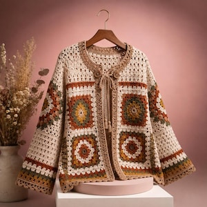 Puede incluir: Un cárdigan de ganchillo con base color crema y motivos de cuadrados de la abuela en tonos amarillo mostaza, verde oliva y naranja quemado. La prenda tiene mangas largas, cuello redondo y un cierre frontal con lazos y borlas.