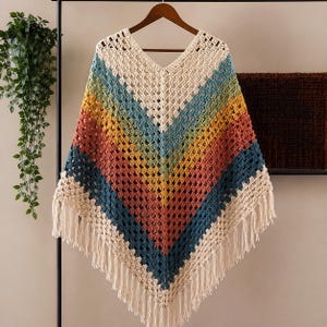 Puede incluir: Un poncho de ganchillo con cuello en V que presenta un patrón de chevrón en crema, azul cerceta, amarillo mostaza y terracota. La prenda está confeccionada con un punto abierto y terminada con largos flecos color crema en el dobladillo.