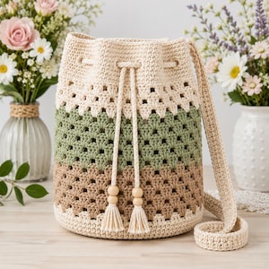 May include: A crochet bucket bag featuring a color-blocked design with horizontal stripes in cream, sage green, and tan. The bag has a drawstring closure with two long cords ending in wooden beads and tassels, and a single wide crochet shoulder strap.