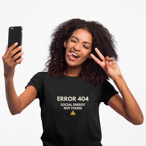May include: A black short-sleeved t-shirt featuring the white text ERROR 404 SOCIAL ENERGY NOT FOUND above a yellow triangle warning icon with an exclamation mark. The shirt is made of a solid black fabric with a crew neckline and short sleeves.