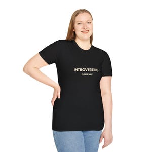 May include: A black short-sleeved t-shirt featuring the text INTROVERTING PLEASE WAIT printed in a light beige color across the chest. The garment has a standard crew neckline and a straight hemline, providing a simple and clean appearance.
