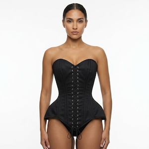 May include: A strapless black corset featuring a structured bodice with vertical paneling and a central front lace-up closure with silver-toned metal eyelets. The garment includes a flared peplum waist detail and a high-cut leg opening design.