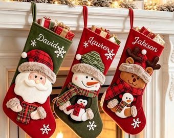 Op de afbeelding: Drie gepersonaliseerde kerstsokken hangen op een rij. De eerste is groen met een kerstman, de tweede rood met een sneeuwpop en pinguïn, en de derde rood met een rendier en sneeuwpop. Elke sok is voorzien van een naam en sneeuwvlokken.