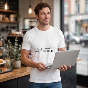 May include: A white short-sleeved t-shirt featuring the black printed text // IT WORKS. DON'T TOUCH IT. The shirt has a crew neckline and is made of a plain fabric, shown here as a casual garment worn with blue denim jeans while holding a laptop.
