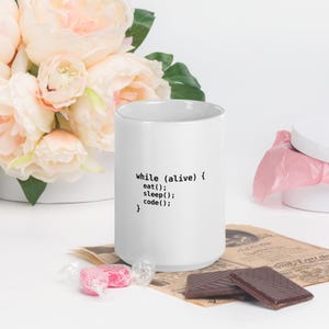 May include: A white ceramic mug featuring black printed text that reads while (alive) { eat(); sleep(); code(); }. The mug is cylindrical with a smooth finish and a simple handle, displaying programming syntax on its front surface.
