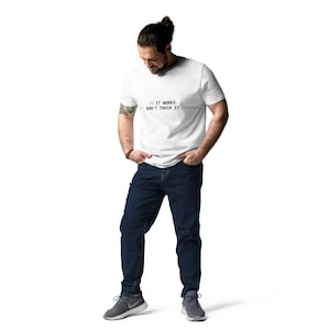 May include: A white short-sleeved t-shirt featuring the black text // IT WORKS. DON'T TOUCH IT. centered on the chest, paired with dark blue denim jeans and grey athletic sneakers. The shirt has a crew neckline and a standard fit design.