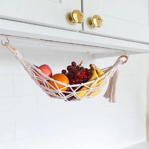 May include: A cream-coloured macrame fruit hammock suspended by two wooden rings from hooks under white kitchen cabinets. The woven net holds a red apple, an orange, a cluster of red grapes, and two yellow bananas, featuring a decorative tassel.