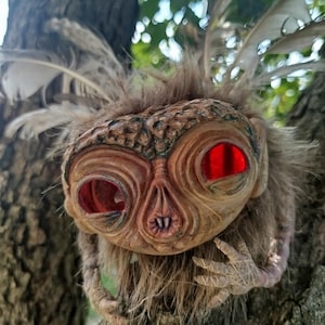 May include: A creature figurine with a textured, tan, wrinkled face and large, bright red, circular eyes. The figure is covered in brown, shaggy faux fur and features long, light-coloured feathers extending from the top of its head.