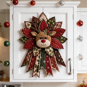 May include: A quilted fabric wall hanging featuring a central reindeer head with a red nose and brown antlers, surrounded by layered, petal-like fabric pieces in red, green, and plaid patterns, accented with holly leaves and berries.