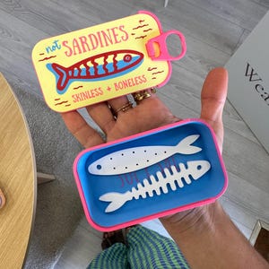Può includere: Un contenitore rettangolare giallo con bordo rosa e coperchio a strappo. Il coperchio presenta una grafica di pesce rosso e blu con il testo "not SARDINES" e "SKINLESS + BONELESS". All'interno del contenitore blu ci sono due oggetti bianchi a forma di pesce.