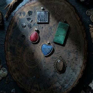 May include: A collection of five pendants featuring textured, scale-patterned inlays in red, blue, green, black, and brown. The pendants are shaped as a square, a teardrop, a heart, a rectangle, and an oval with an ornate metal frame.