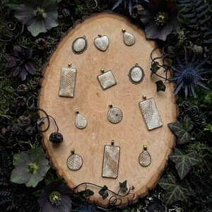 May include: A collection of eleven pendants featuring a textured, cream-colored snakeskin-like pattern set in antique bronze-toned metal frames. The pendants vary in shape, including rectangles, circles, ovals, hearts, and teardrops, all arranged on a wood slice.