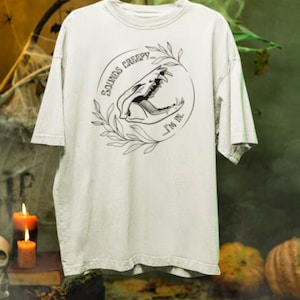 May include: A light-coloured short-sleeve t-shirt featuring a black line-art graphic of an animal skull inside a circular frame of leaves. The text "SOUNDS CREEPY... ...I'M IN." is printed in a curved font around the skull illustration.