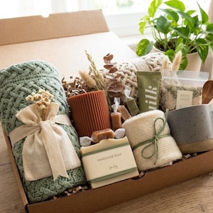 May include: A gift box containing a sage green cable knit blanket, a ribbed terracotta candle, a bar of handmade soap, a beige knit cloth, a speckled gray mug, a tube of milk lotion, a bag of bath salts, dried flowers, and small wrapped candies.