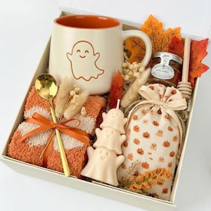 Könnte beinhalten: Eine Geschenkbox mit einer cremefarbenen Tasse mit orangefarbenem Innenraum und Geistermotiv, einem goldenen Löffel, einer flauschigen orange-weißen Decke, drei gestapelten Geisterkerzen, einem Honigglas mit Holzlöffel und einem Beutel.