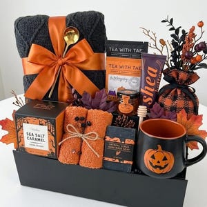 May include: A gift basket containing a dark gray knit blanket with an orange ribbon, a gold spoon, Tea With Tae tea, a jar of honey, a shea butter tube, orange towels, oatmeal soap, a black mug with an orange pumpkin, and decorative autumn leaves.
