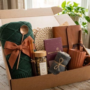 May include: A cardboard gift box containing a dark green cable-knit blanket with a rust-colored ribbon, a copper spoon, amber glass straws, a jar of herbal tea, a round woven coaster, a Moonstruck chocolate bar, a brown notebook, and a mug.