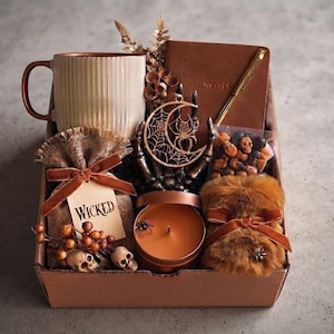 Op de afbeelding: Een geschenkdoos met een geribbelde beige mok, een bruin notitieboek met een gouden pen, een houten ornament van een halve maan met een spin, een zakje met het label WICKED met schedeltjes, een koperkleurige kaars en een pluizig bruin tasje.
