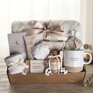 May include: A gift basket containing a beige knit blanket with a satin ribbon, a Creekside Ceramics mug, a jar of Serenity Salts, a bar of Lavender & Bergamot soap, a Sheer Botanica tube, a white flower, a small pouch, and a Cloud Cushions item.