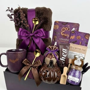 May include: A gift set featuring a dark brown faux fur blanket tied with a purple ribbon, a purple ribbed mug with a bat graphic, a brown pumpkin-shaped ceramic jar, a jar of purple bath salts, a tea packet, a face mask, and two gold-tone spoons.
