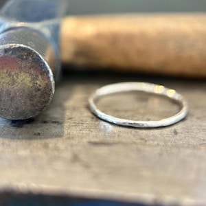 May include: A thin, circular silver ring with a textured, hammered surface finish. The ring is positioned on a flat, metallic surface next to a metal hammer with a wooden handle, showcasing the reflective quality of the silver metal band.