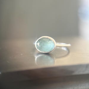 May include: A silver ring featuring a faceted, oval-shaped, pale mint green gemstone set in a smooth metal bezel. The ring band is thin and polished, reflecting light on its surface, showcasing a clear, translucent stone with subtle internal facets.