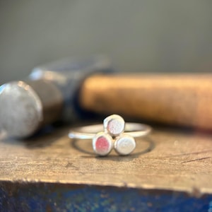 May include: A silver ring featuring three small, circular, textured metal discs arranged in a cluster on the band. The ring has a matte finish and is positioned in front of a blurred metal hammer, highlighting the handcrafted nature of the jewelry piece.