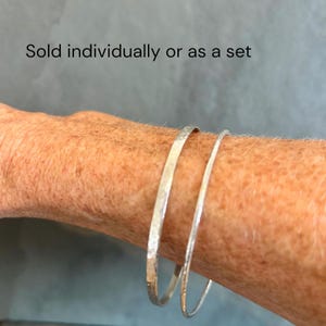 May include: Two silver-coloured bangles with a hammered texture. One bangle is slightly thicker than the other. The text "Sold individually or as a set" is visible above the bracelets. The bangles have a metallic finish and a circular shape.
