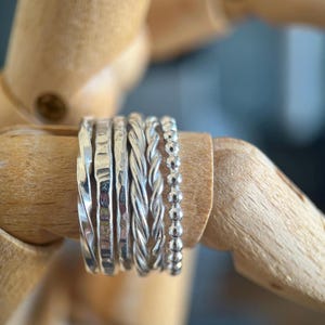 May include: A set of six silver rings featuring varied textures, including smooth twisted bands, hammered surfaces, and a delicate beaded design. The collection consists of distinct metal bands arranged side by side, showcasing reflective surfaces and intricate detailing.