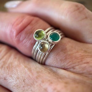 May include: A stack of silver rings featuring three round, bezel-set cabochon gemstones in shades of light green, dark green, and a translucent amber tone. The bands are smooth and metallic, creating a layered appearance on the finger.