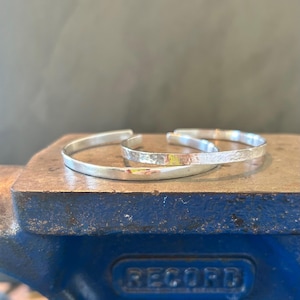 May include: Two silver-colored metal cuff bracelets. One bracelet features a smooth, polished surface, while the other bracelet has a textured, hammered finish. Both bracelets are open-ended and circular in shape, displaying a metallic sheen.