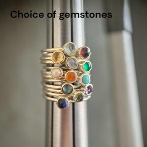 May include: A stack of silver rings on a metal mandrel, each featuring a different gemstone. The text 'Choice of gemstones' is visible at the top. The stones include red, green, blue, orange, purple, yellow, white, and iridescent varieties.