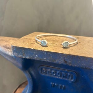 May include: A silver cuff bracelet featuring two small, irregular, light blue-green stones at each open end. The bracelet has a smooth, matte finish and is resting on the flat, metallic surface of a blue anvil with the word RECORD embossed on it.