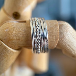 May include: A set of five stacked silver rings featuring varied textures, including a twisted band, a beaded band, and three smooth bands with different finishes, all worn together as a single cohesive piece of jewellery on a wooden mannequin hand.