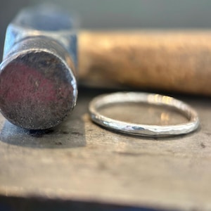 Könnte beinhalten: Ein dünner, silberfarbener Ring mit einer texturierten, gehämmerten Oberfläche. Der Ring liegt neben dem abgerundeten Metallkopf eines Werkzeugs mit Holzgriff und zeigt den metallischen Glanz sowie das unebene, handgefertigte Erscheinungsbild.